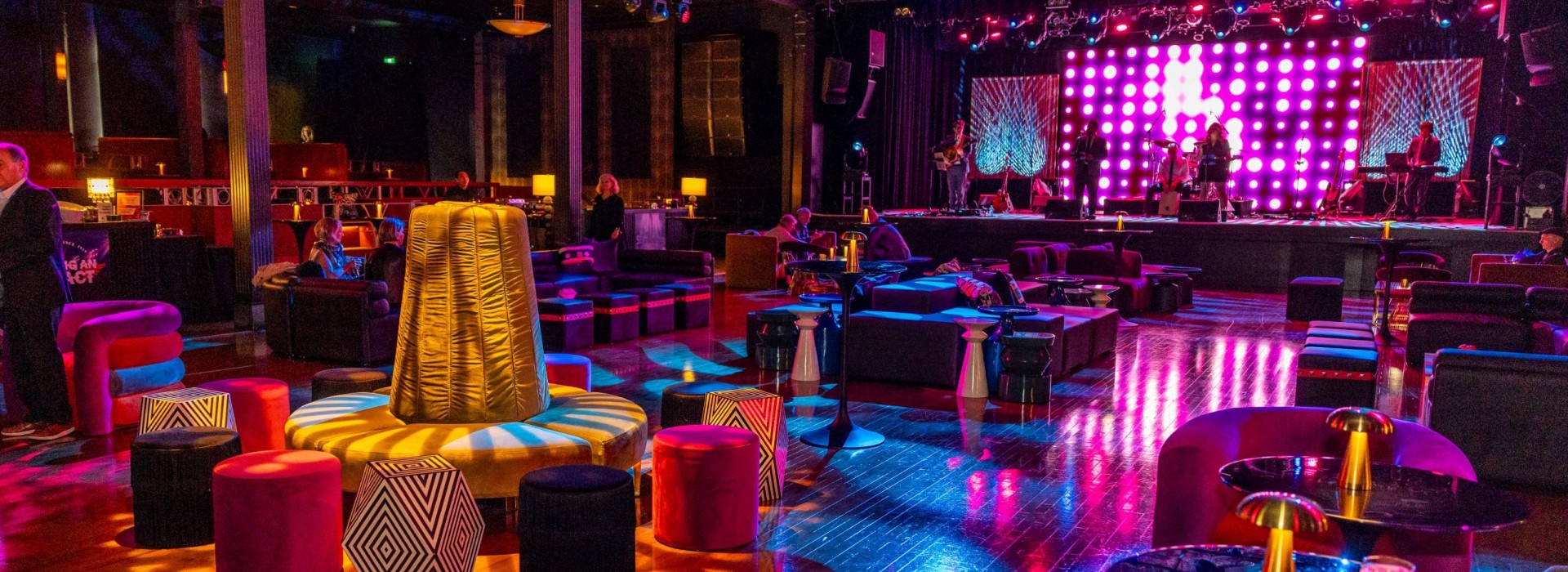 A stylish, dimly lit event space with colourful seating, a stage illuminated by pink and purple lights, and a few attendees seated around small tables.